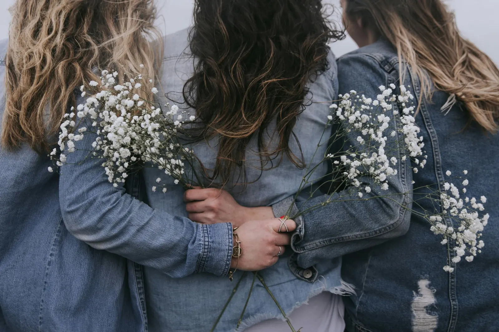 Three women seen from behind, arms around each other, holding small white flowers